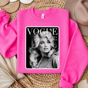 Può includere: Una felpa girocollo rosa acceso con una stampa grafica rettangolare sul davanti. La stampa è un ritratto in bianco e nero di una donna con voluminosi capelli biondi, con il testo VOGUE e DOLLY EDITION stampato in bianco.