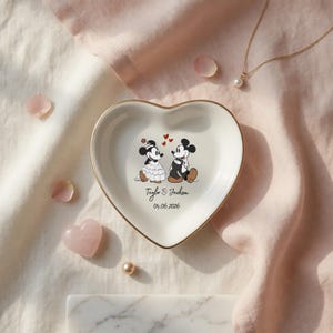 May include: A white heart-shaped ceramic trinket dish with a gold-toned rim. The center features a graphic of two cartoon mice in formal wedding attire, three small red hearts, and the text Taylor & Jackson followed by the date 04.06.2026.