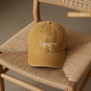 May include: A mustard yellow baseball cap featuring a white line art embroidery of a sun rising over wavy water. The design is centered on the front panels of the cap, which has a structured crown, a curved brim, and a button on the top center.