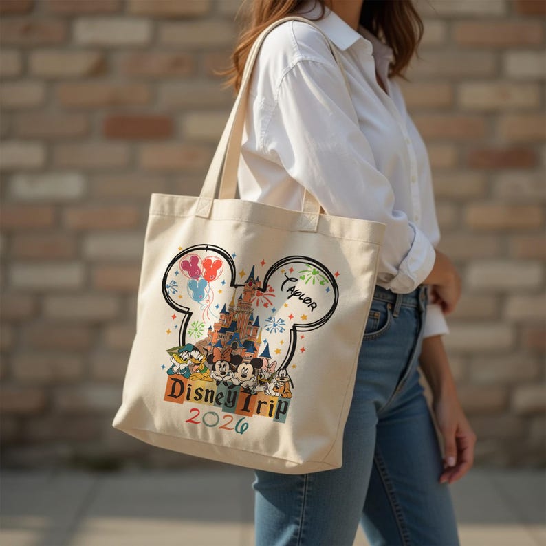 Pu&ograve; includere: Una borsa tote in tela color crema con la grafica di un castello all'interno di una silhouette a orecchie di Topolino, circondata da personaggi dei cartoni animati, palloncini colorati e fuochi d'artificio. Il testo recita Disney Trip 2026 con il nome Taylor.