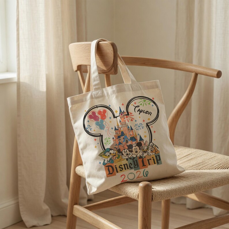 Pu&ograve; includere: Una borsa in tela color crema con un grande contorno nero a forma di orecchie di Topolino. All'interno si trova un'illustrazione colorata di un castello, fuochi d'artificio e personaggi. Il testo riporta Taylor, Disney Trip e l'anno 2026.