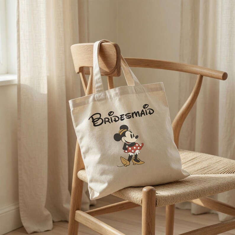 May include: A cream-colored canvas tote bag featuring the word Bridesmaid in black script lettering above a graphic of Minnie Mouse wearing a red and white polka dot skirt, a yellow hat, and yellow shoes, with two top handles for carrying.