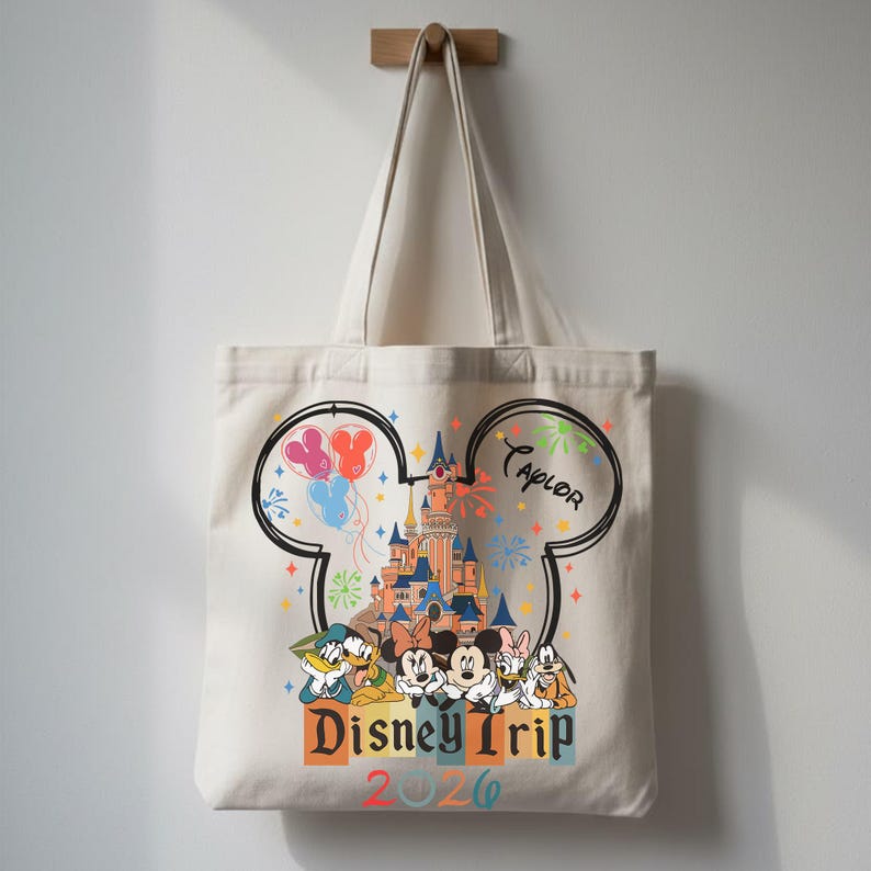 Pu&ograve; includere: Una borsa in tela color crema con la grafica di un castello all'interno di una sagoma a orecchie di Topolino. Il design include personaggi, palloncini colorati, fuochi d'artificio, il testo "Disney Trip 2026" e il nome "Taylor" in corsivo.