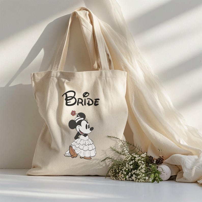 May include: A cream-colored canvas tote bag featuring the word BRIDE in black lettering above a graphic of a cartoon mouse character wearing a ruffled white dress and a small flower on its head, with two long shoulder straps for carrying.