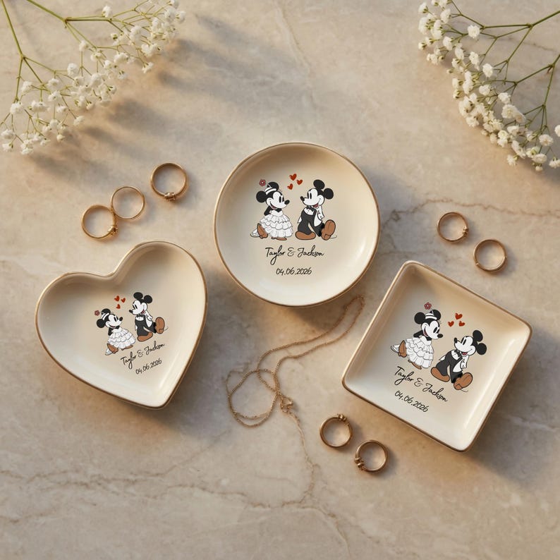 May include: Three cream-colored ceramic trinket dishes in heart, round, and square shapes. Each dish features a graphic of Mickey and Minnie Mouse, the text Taylor & Jackson, and the date 04.06.2026. Gold-toned rims accent the edges of each dish.