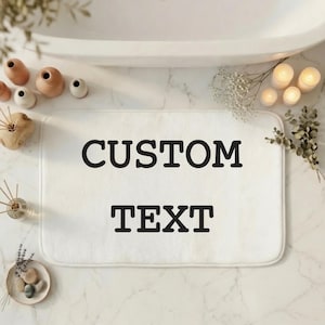 May include: A rectangular white bath mat featuring the words CUSTOM TEXT printed in a bold black sans-serif font. The mat has rounded corners and a soft, plush texture, positioned centrally as a functional bathroom accessory for floor use.
