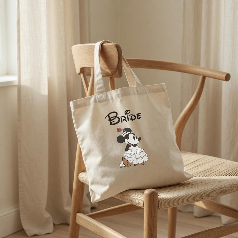 May include: A cream-colored canvas tote bag featuring the word Bride in black script lettering above a graphic of Minnie Mouse wearing a ruffled white dress and a small hat with a flower. The bag has two fabric handles and a rectangular shape.