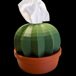 Peut inclure: Un cache-boîte à mouchoirs vert en forme de cactus à facettes, placé dans un pot rond de couleur terre cuite. Un mouchoir blanc unique est tiré par l'ouverture supérieure du cactus, s'étendant vers le haut depuis le centre de la structure côtelée.