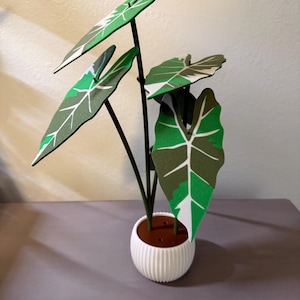 May include: A decorative artificial plant with large green leaves featuring white and dark green patterns. The plant has a dark stem and is potted in a white, ribbed ceramic pot. The plant is a decorative item.