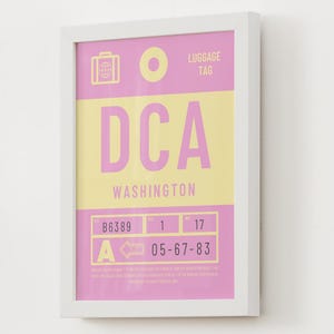 May include: A framed print featuring a stylized airport luggage tag design in pink and yellow. The text reads DCA, WASHINGTON, LUGGAGE TAG, B6389, 1, 17, A, and 05-67-83. The design includes a suitcase icon and a circular hole graphic.