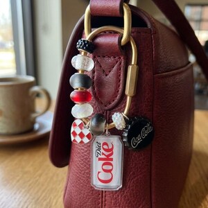 May include: A gold-toned metal carabiner keychain attached to a dark red leather handbag. The keychain features various beads in black, white, and red, a red and white checkered heart charm, a Diet Coke can charm, and a black Coca-Cola bottle cap.