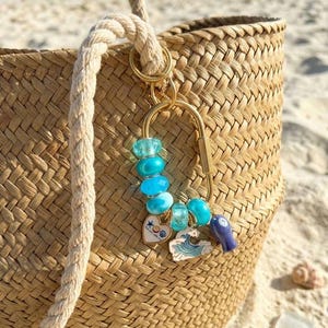 May include: A woven straw tote bag with thick rope handles featuring a gold-toned carabiner keychain. The keychain is adorned with various blue glass beads, a heart-shaped charm, a wave-shaped charm, and a small blue fish-shaped pendant.