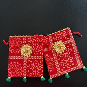 May include: Two rectangular red fabric drawstring pouches featuring a repeating geometric pattern in yellow and orange. Each pouch is adorned with a central gold-colored metallic flower embellishment, gold trim at the top, and three small green pom-poms.