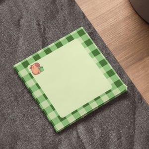 May include: A pad of square sticky notes. The notes have a light green background and a green and white checked border. A small cartoon illustration of a brown animal holding a green cup is in the corner.
