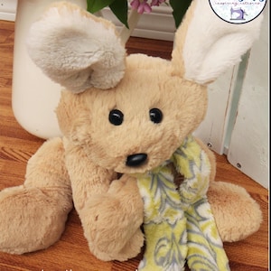 May include: A tan and white plush toy rabbit wearing a green and gray scarf. The rabbit is sitting on a wooden surface. The text "Azalea the Snowshoe Hare Stuffed Animal Sewing Pattern" is printed below the rabbit.