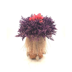May include: A woven basket container filled with a dense arrangement of deep purple statice flowers and a cluster of bright pink blossoms. The basket is adorned with a fringe of tan dried grasses and a silver chain featuring a light-colored crystal pendant.