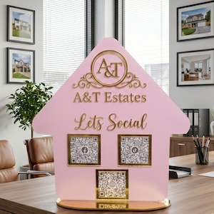 May include: A pink house-shaped sign with gold accents and the text "A&T Estates Lets Social." The sign has three QR codes and sits on a wooden desk in an office setting. Framed photos of houses are on the wall.