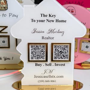 May include: A white acrylic house-shaped sign with gold accents. The sign features the text "The Key To your New Home" and "Jessica Martinez Realtor". Two QR codes are displayed below the text, with the words "Buy. Sell. Invest."