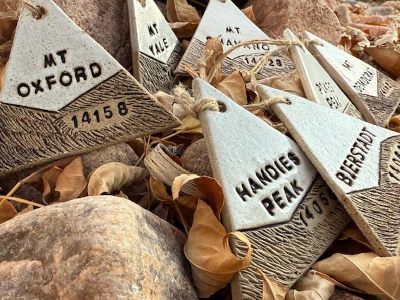 Op de afbeelding: Driehoekige keramische ornamenten met namen van bergtoppen en hoogtes, waaronder "MT OXFORD 14158" en "HANDIES PEAK 1405". De ornamenten zijn wit met bruine, getextureerde bases en zijn geregen met touw.