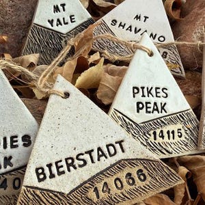 Op de afbeelding: Driehoekige keramische ornamenten met bergnamen en hoogtes, waaronder "MT YALE", "MT SHAVANO", "PIKES PEAK" en "BIERSTADT". Elk ornament heeft een getextureerd, donkerbruin onderste gedeelte en een cr&egrave;mekleurig bovenste gedeelte, geregen met touw.