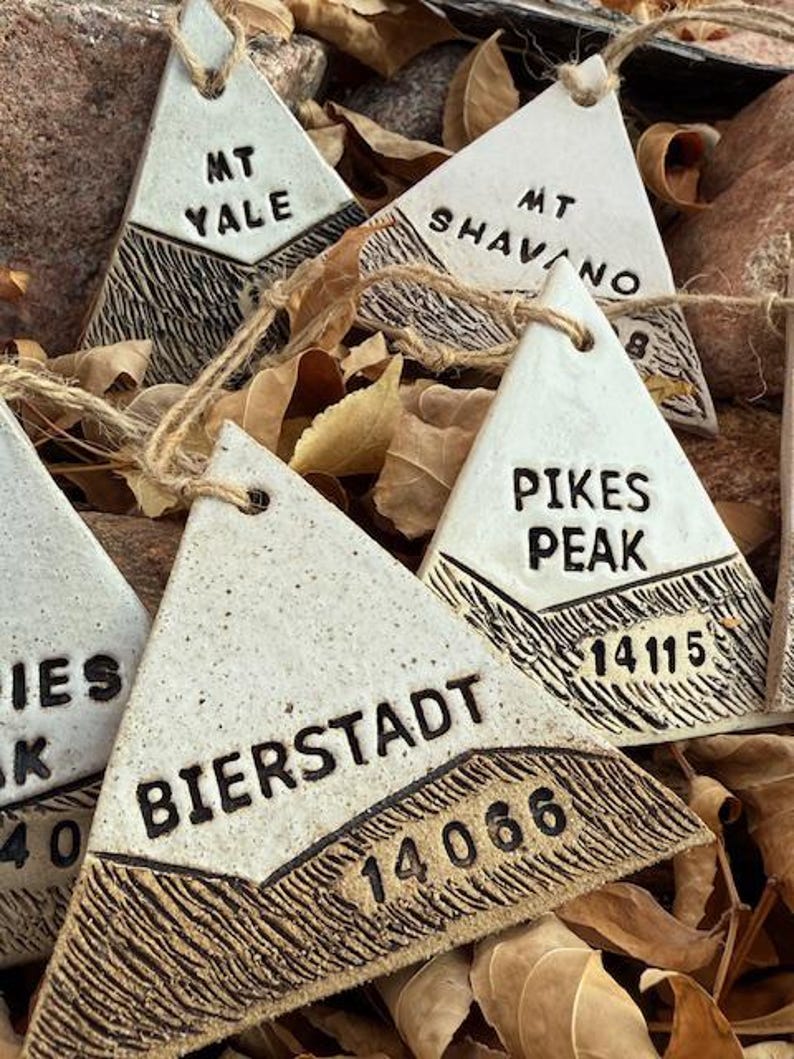 Op de afbeelding: Driehoekige keramische ornamenten met bergnamen en hoogtes, waaronder "MT YALE", "MT SHAVANO", "PIKES PEAK" en "BIERSTADT". Elk ornament heeft een getextureerd, donkerbruin onderste gedeelte en een cr&egrave;mekleurig bovenste gedeelte, geregen met touw.