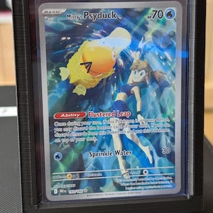 以下が含まれることがあります： 黒いプラスチック製のディスプレイフレームに入ったMisty's Psyduckのトレーディングカード。カードには黄色いコダックと青い服を着たキャラクターが青い水背景の中に描かれており、Flustered LeapとSprinkle Waterというテキストが確認できます。