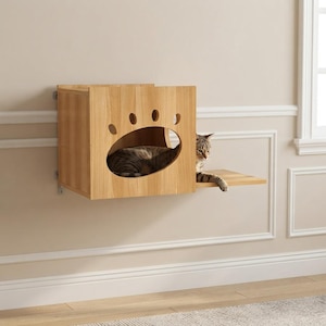 May include: A light-colored wooden wall-mounted cat house featuring a paw-shaped cutout entrance. The structure includes an attached horizontal shelf extending from the side, with a tabby cat resting inside the enclosure and resting a paw on the shelf.