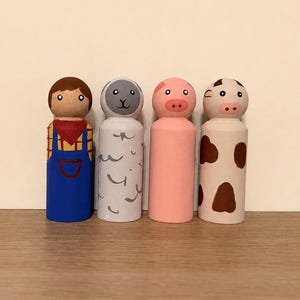 May include: A set of four wooden peg dolls featuring a farmer in blue overalls, a grey sheep with wool texture markings, a solid pink pig, and a white cow with brown spots. Each figure has a rounded head with painted facial features.