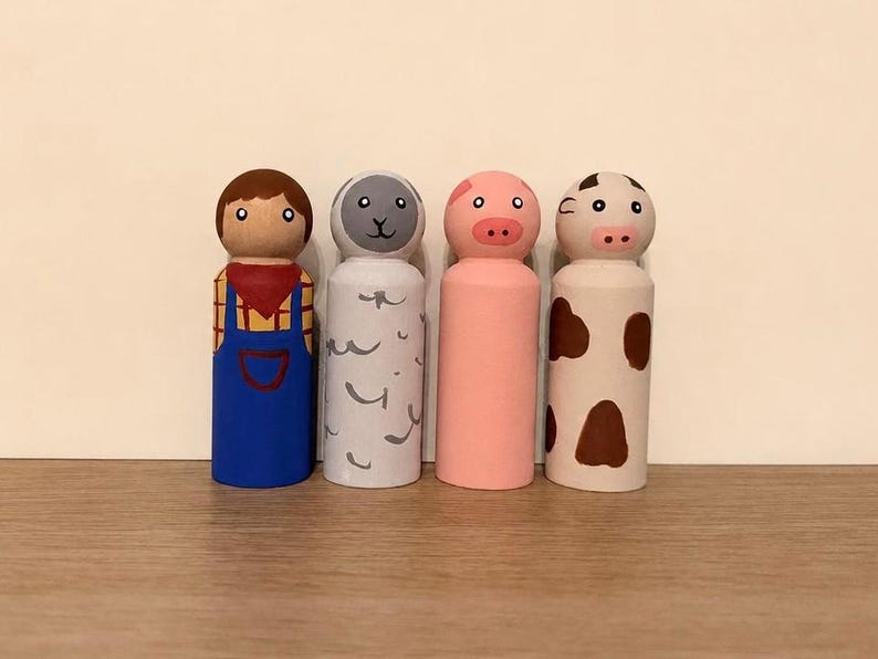 May include: A set of four wooden peg dolls featuring a farmer in blue overalls, a grey sheep with wool texture markings, a solid pink pig, and a white cow with brown spots. Each figure has a rounded head with painted facial features.