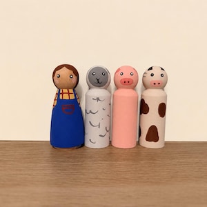 May include: Four hand-painted wooden peg dolls representing a farm scene. The set includes a figure in a blue apron, a grey sheep with curly markings, a solid pink pig, and a white cow with brown spots. Each figure has a smooth, rounded top.