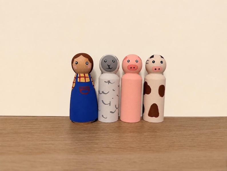 May include: Four hand-painted wooden peg dolls representing a farm scene. The set includes a figure in a blue apron, a grey sheep with curly markings, a solid pink pig, and a white cow with brown spots. Each figure has a smooth, rounded top.