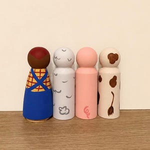 May include: Four wooden peg dolls standing in a row. The first has a brown head, yellow plaid shirt, and blue overalls. The second is light grey with small grey cloud shapes. The third is solid light pink. The fourth is cream with brown spots.