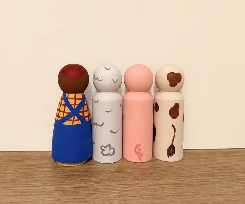 May include: Four wooden peg dolls standing in a row. The first has a brown head, yellow plaid shirt, and blue overalls. The second is light grey with small grey cloud shapes. The third is solid light pink. The fourth is cream with brown spots.