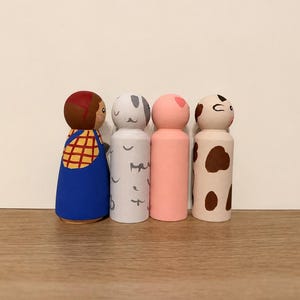 May include: Four wooden peg dolls standing in a row. The first is painted with brown hair, a yellow plaid shirt, and blue overalls. The second is white with grey markings. The third is solid light pink. The fourth is cream with brown spots.