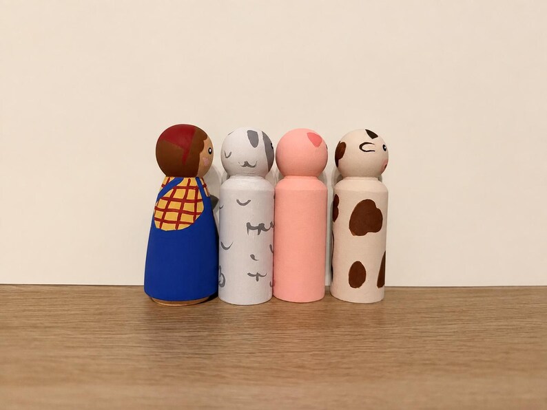 May include: Four wooden peg dolls standing in a row. The first is painted with brown hair, a yellow plaid shirt, and blue overalls. The second is white with grey markings. The third is solid light pink. The fourth is cream with brown spots.