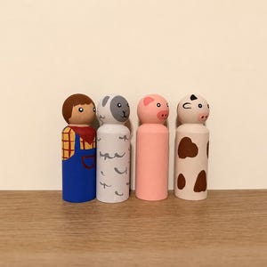 May include: A set of four hand-painted wooden peg dolls featuring a farmer in blue overalls and a red scarf, a grey sheep with white markings, a solid pink pig, and a white cow with brown spots, all standing in a row on a flat surface.