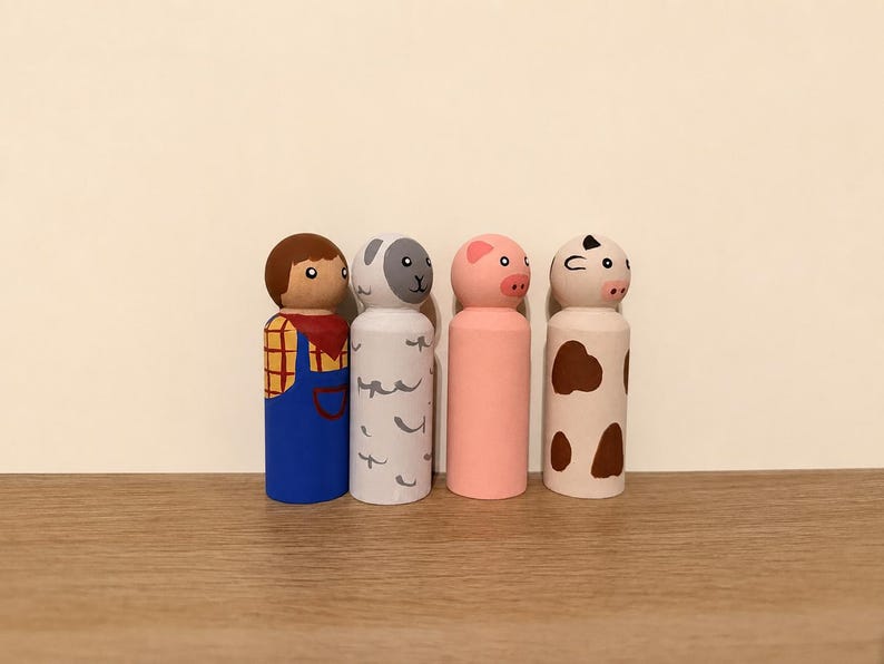May include: A set of four hand-painted wooden peg dolls featuring a farmer in blue overalls and a red scarf, a grey sheep with white markings, a solid pink pig, and a white cow with brown spots, all standing in a row on a flat surface.