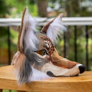 May include: A detailed, handmade wolf mask with brown, tan, and white fur. The mask features a realistic design with a black nose, detailed eyes, and fluffy white fur around the ears. The mask is placed on a wooden surface.