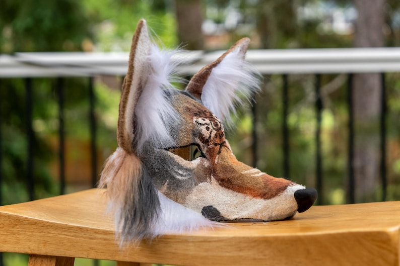 May include: A detailed, handmade wolf mask with brown, tan, and white fur. The mask features a realistic design with a black nose, detailed eyes, and fluffy white fur around the ears. The mask is placed on a wooden surface.