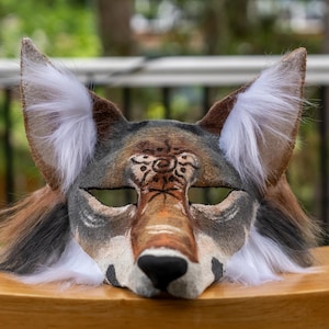 May include: A handcrafted coyote mask with a detailed design. The mask features a brown, gray, and white color scheme, with a prominent snout and large, pointed ears. The mask is made of felt and faux fur.