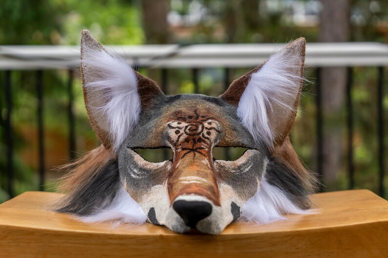 May include: A handcrafted coyote mask with a detailed design. The mask features a brown, gray, and white color scheme, with a prominent snout and large, pointed ears. The mask is made of felt and faux fur.