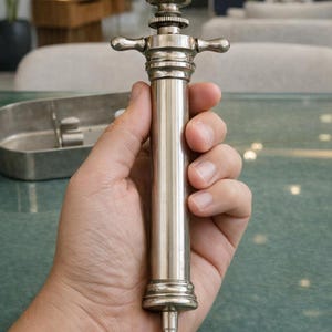 May include: A vintage-style silver-toned metal syringe featuring a cylindrical barrel, a plunger mechanism with a circular top handle, and two side-extending finger grips. The device has a tapered nozzle at the base and a polished metallic finish.