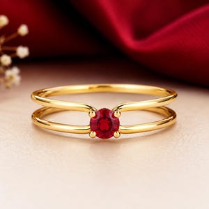 Könnte beinhalten: Ein goldener Ring mit einem runden, tiefroten Edelstein in einer Vier-Krappen-Fassung. Die Ringschiene ist im Split-Shank-Design gestaltet, bei dem sich zwei dünne, glatte Golddrähte teilen und an der zentralen Fassung wieder treffen.