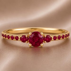 Op de afbeelding: Een gouden ring met een prominente, rondgeslepen rode robijn in het midden, geflankeerd door twee kleinere ronde robijnen aan weerszijden, met een reeks kleine rode robijnen ingelegd langs de dunne, gepolijste gouden band.