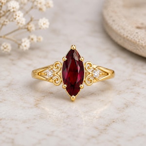 Op de afbeelding: Een gouden ring met een centrale dieprode edelsteen in marquise-slijpvorm, vastgehouden door zes pootjes. De ringband vertoont aan beide kanten ingewikkelde krulpatronen, elk geaccentueerd met een kleine, ronde, heldere witte edelsteen.