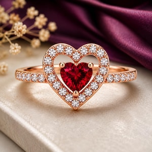 May include: A rose gold ring featuring a central heart-shaped red gemstone surrounded by a halo of small round white stones. The band is also set with a row of small round white stones, creating a continuous sparkling effect along the metal shank.