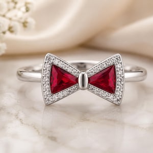 May include: A silver-toned ring featuring a bow-shaped centerpiece. The bow is composed of two triangular-cut deep red stones, each framed by a border of small, clear, sparkling stones, with a smooth silver center accent connecting the two sides.