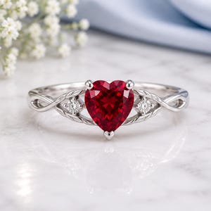 Peut inclure: Une bague argentée ornée d'une pierre précieuse rouge foncé en forme de cœur au centre. L'anneau présente un motif métallique torsadé complexe de chaque côté, agrémenté d'une petite pierre claire ronde sertie sur chaque épaulement.