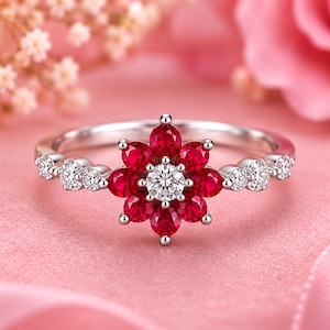 Peut inclure: Une bague en argent avec un motif floral central composé de pierres rouges taille poire entourant une pierre blanche ronde. L'anneau est orné de trois petites pierres blanches rondes de chaque côté, sur une finition en métal argenté poli.
