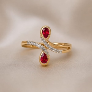 May include: A gold ring featuring two pear-shaped red gemstones set at opposite ends of a bypass-style band. The band is accented with small, clear pavé-set stones along the curved arms that extend from the central ruby-colored teardrop settings.